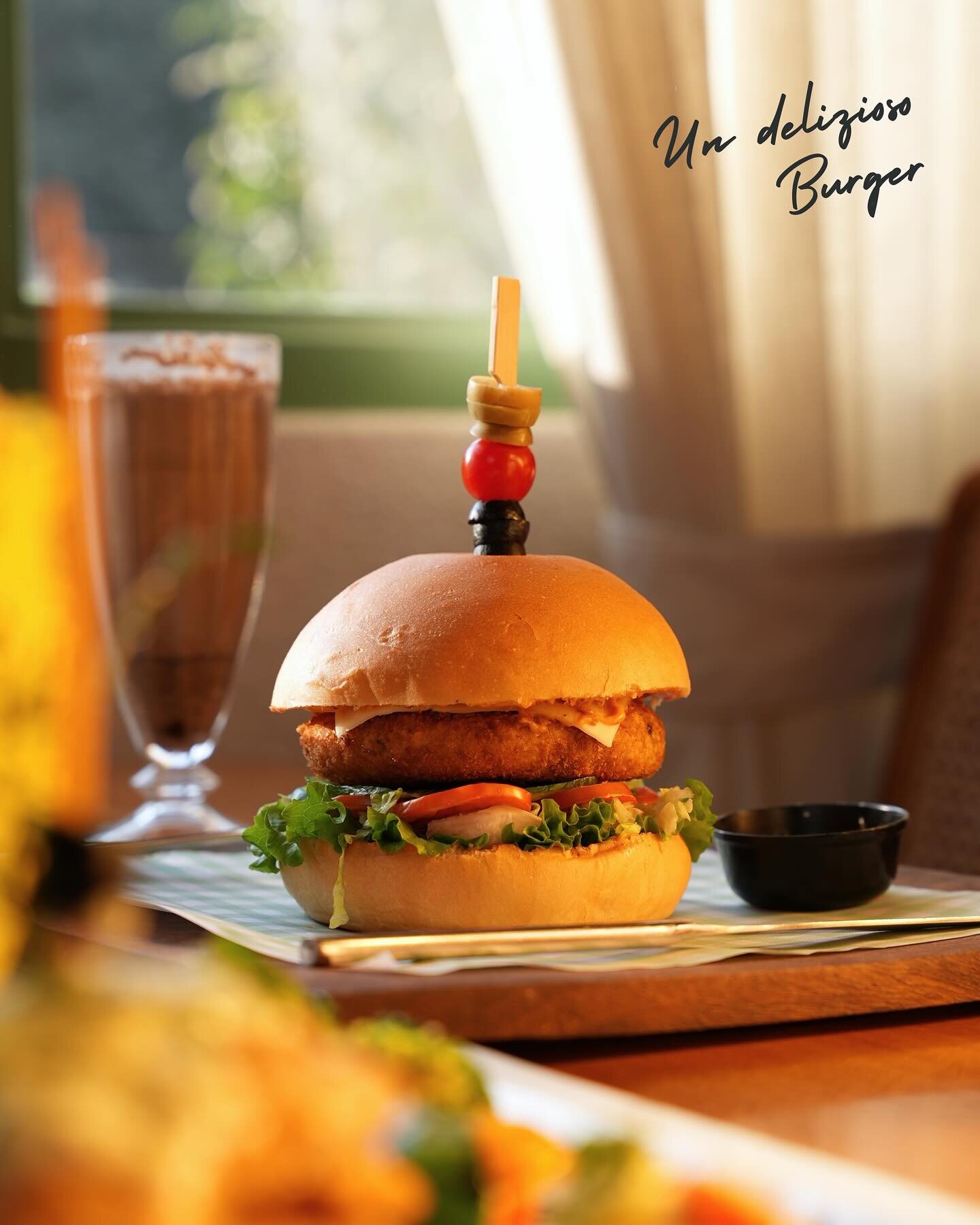 A burger in a brioche bun beside a tall iced drink, in afternoon light